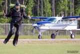 A person in a black jumpsuit is landing with a parachute on a grassy area. A white and blue airplane is parked in the background near a line of trees.