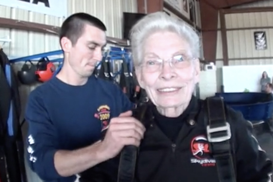 An elderly woman with white hair, smiling and wearing a skydiving jumpsuit with harnesses, stands indoors with a younger man adjusting her gear. The setting appears to be a skydiving facility.