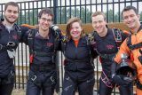 A group of five people in skydiving gear stand together, smiling for a photo. They are wearing black jumpsuits with harnesses. A fence and trees are visible in the background.