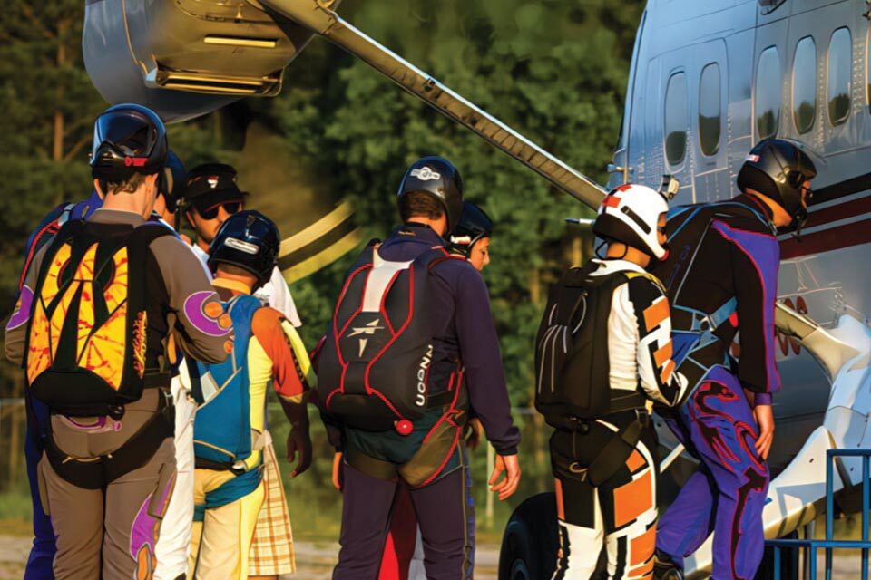 A group of skydivers in colorful jumpsuits and helmets prepare to board a small aircraft. They are lined up beside the plane, ready for a skydiving adventure, with parachute backpacks visible. The scene is set in the late afternoon sunlight.