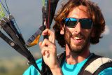 A man with shoulder-length brown hair and a beard smiles widely while wearing sunglasses and a blue shirt. He holds the cords of a paraglider, wearing a harness, with a clear sky in the background.