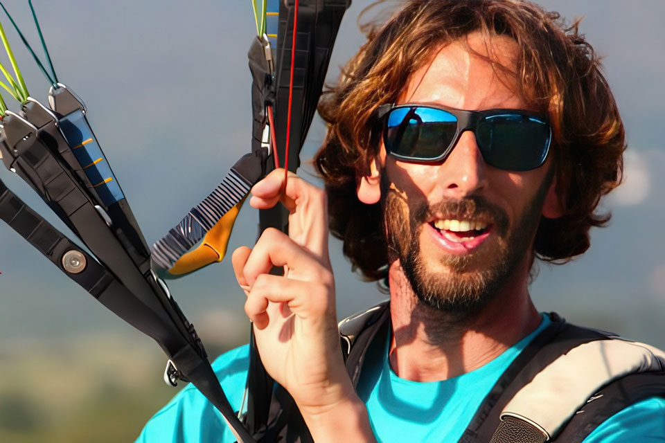 A man with shoulder-length brown hair and a beard smiles widely while wearing sunglasses and a blue shirt. He holds the cords of a paraglider, wearing a harness, with a clear sky in the background.