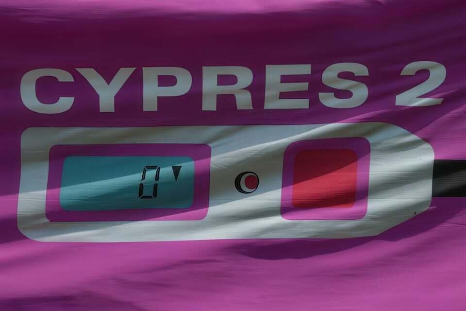 Close-up of a banner displaying a digital screen with "CYPres 2" written above it. The screen shows a number zero and a button is visible beside it. The background is mainly pink and purple with shadows.