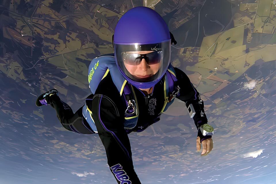 A skydiver in a blue and black suit with matching helmet free-falls through the sky. The ground is visible far below, and the sky is clear, creating a sense of speed and freedom.