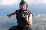 A skydiver wearing a helmet and goggles floats in the air, making a hand gesture, with fields and a body of water visible far below. He is equipped with a parachute, and the sky is partly cloudy.