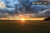 A vast open field at sunset with dramatic clouds in the sky. The sun is setting near the horizon, casting a warm glow across the landscape. A sign in the top right corner reads "New Heights Club.