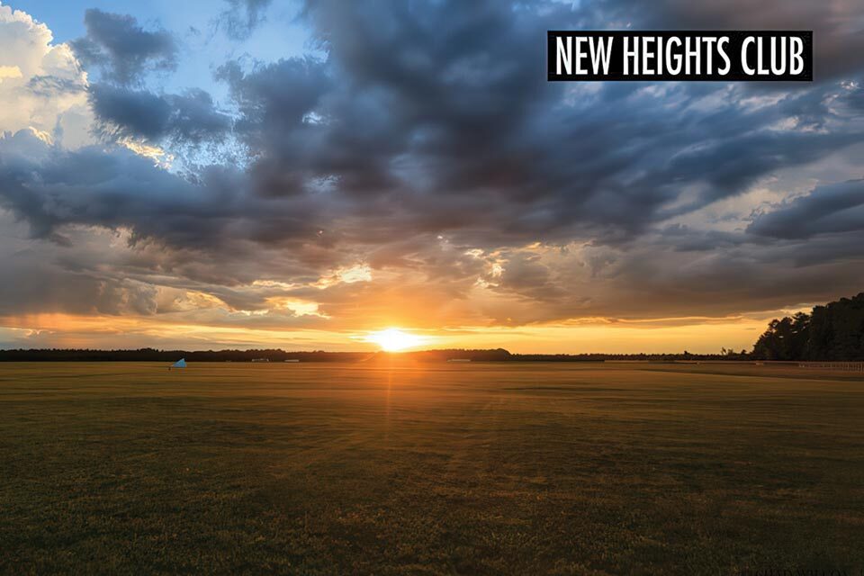 A vast open field at sunset with dramatic clouds in the sky. The sun is setting near the horizon, casting a warm glow across the landscape. A sign in the top right corner reads "New Heights Club.