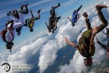 A group of skydivers in a freefall formation above the clouds, wearing colorful jumpsuits. The sky is clear and blue, offering a stunning aerial view.