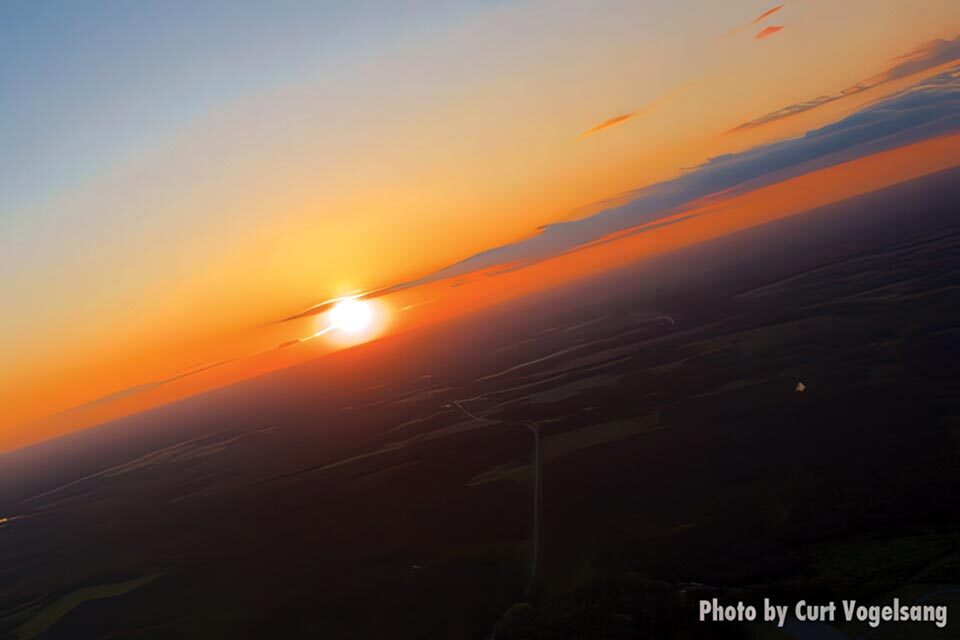 Aerial view of a sunset over a vast landscape with fields and winding roads. The sun is near the horizon, casting a warm orange glow across the sky. Dark clouds hover above the horizon.