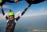 A skydiver wearing a bright green helmet and black jumpsuit is smiling mid-air, while holding onto another person. The sky is clear and the landscape below features fields and farmland.