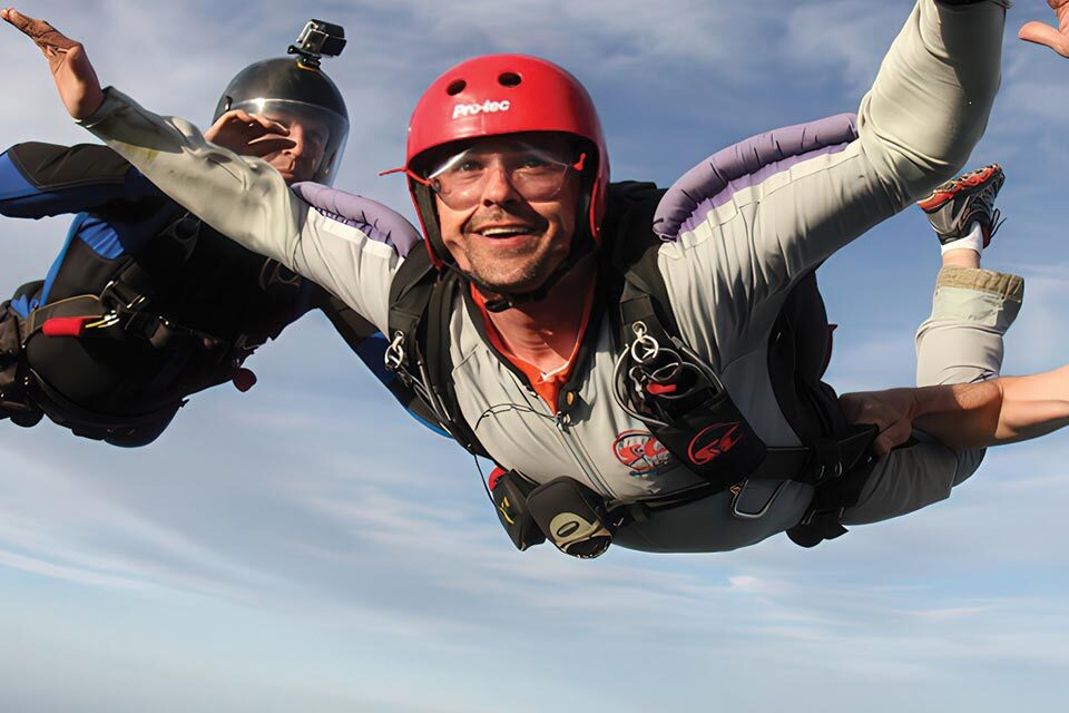 Two skydivers are in free fall against a clear blue sky. The man in the foreground wears a red helmet and a gray jumpsuit, smiling at the camera. The second skydiver wears a black helmet, partially visible, and appears focused.