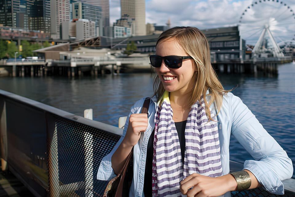 A woman wearing sunglasses and a striped scarf smiles while standing on a pier with a city skyline and a Ferris wheel in the background.