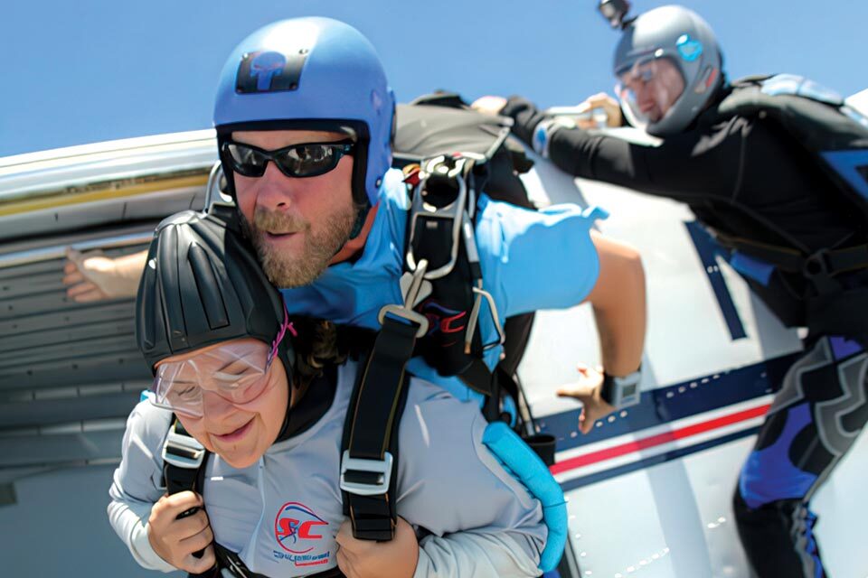 A tandem skydiver with an instructor jumps out of an airplane. Both are wearing helmets and protective gear, while another person is visible inside the plane. The sky is clear and blue.