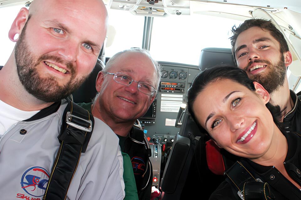 Four people are smiling and sitting inside a plane cockpit, wearing harnesses. The cockpit is equipped with various controls and displays, with daylight visible through the windows.