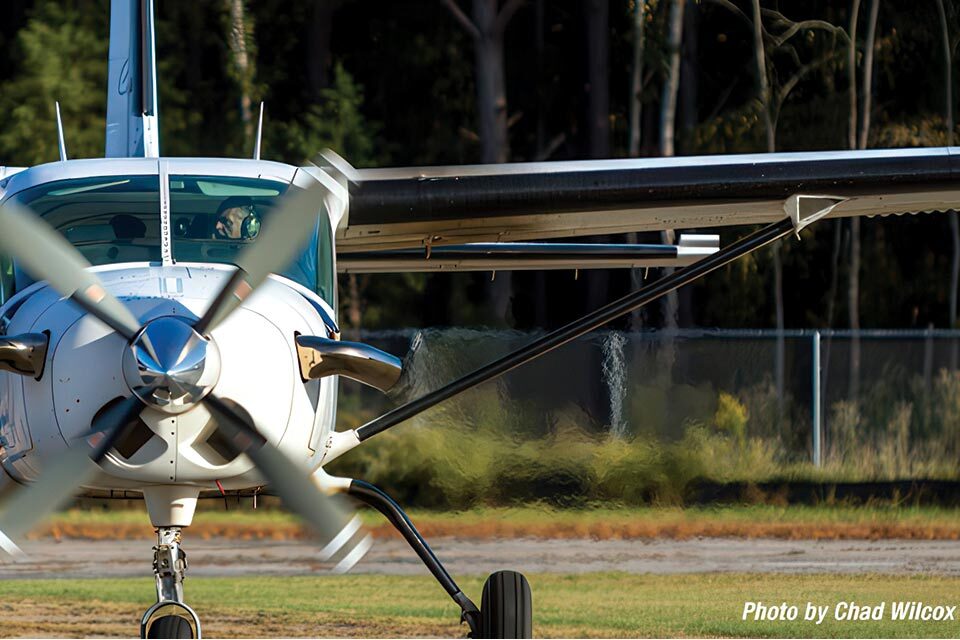 A small propeller plane on a grassy airfield is captured mid-motion with blurred spinning propeller blades. The background features trees and a wire fence. (Photo by Chad Wilcox).