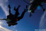 Two people tandem skydiving, silhouetted against a bright sun and clear blue sky. The airplane is seen above them, with glowing navigation lights, as they freefall. Photo credit: Quincy Kennedy.