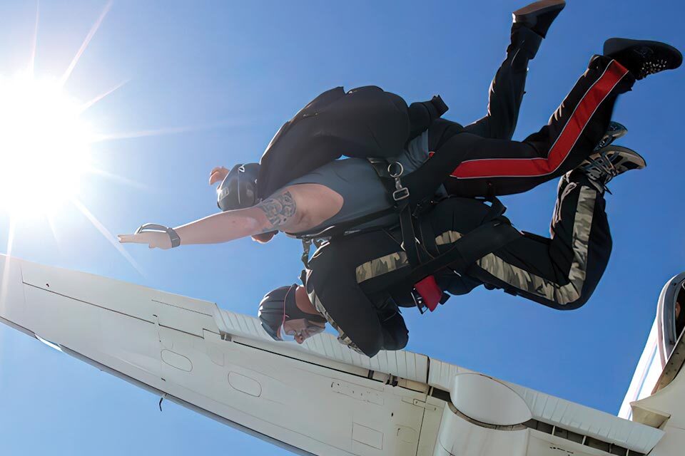 Two skydivers are tandem jumping from an airplane. The instructor is strapped to the student, guiding the fall. They are free-falling against a clear blue sky with the sun shining brightly above.