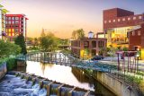 Scenic view of a cityscape at sunset featuring a modern bridge over a calm river with cascading steps, surrounded by trees and vibrant buildings. A clear sky and soft lighting create a peaceful atmosphere.