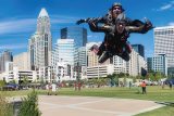 Two people in skydiving suits are mid-air over a cityscape on a clear day. They appear to be floating above a park with green grass and scattered people below. Tall buildings and a bright blue sky form the backdrop.