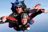 Two people are tandem skydiving. The instructor behind is smiling and giving a thumbs-up, while the person in front looks exhilarated. Both are wearing helmets and goggles against a clear blue sky.