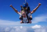 A person skydiving in tandem with an instructor, smiling and giving a thumbs-up. They are wearing harnesses and helmets, with a clear blue sky and clouds in the background.