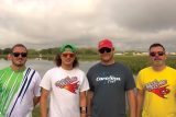 Four men stand together outdoors in front of a lake. They are wearing casual T-shirts, sunglasses, and caps. The sky is cloudy, and there is greenery in the background.