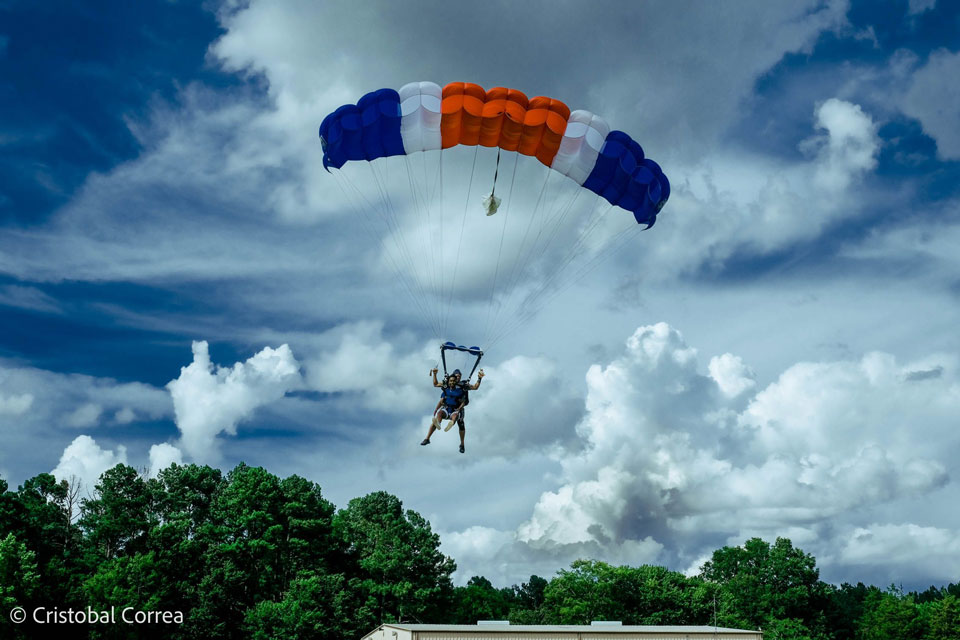 A person is paragliding with a blue, white, and orange parachute against a backdrop of a cloudy sky. Green trees and a white building are visible below.