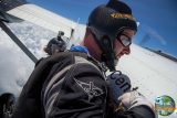 A skydiver wearing a helmet and goggles stands at the open door of an airplane, preparing to jump. Another skydiver is visible behind, ready for the dive. The sky is clear with some clouds.