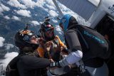 Three skydivers with helmets and gear prepare to jump out of a plane. The skydivers, two in light blue and one in orange, are holding onto each other, and the ground is visible far below through scattered clouds.