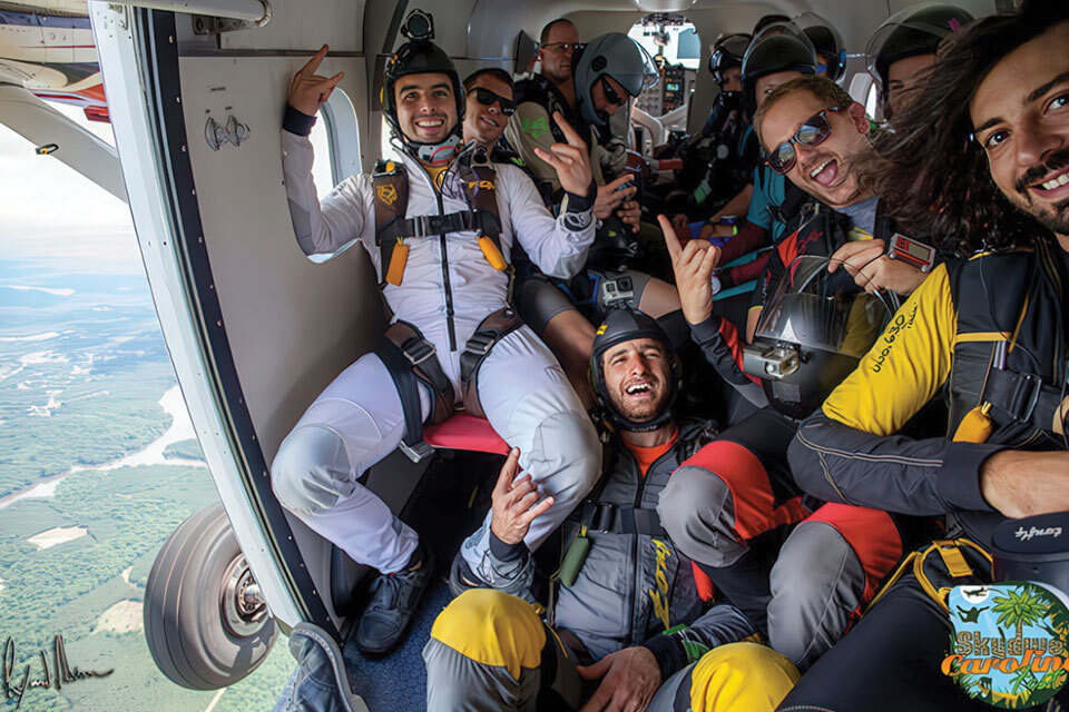 A group of skydivers in colorful jumpsuits are smiling and posing inside an open airplane, ready to jump. The airplane door is open, revealing a scenic view of the landscape below. Everyone looks excited and is making hand gestures.