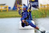 A person in blue skydiving gear is captured mid-landing, skimming the surface of a body of water. The skydiver wears a helmet with a camera attached. The background features a grassy area and some structures.