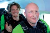 Two men in a plane wearing skydiving gear smile at the camera. The man in front is bald, while the man behind has curly hair and gives a thumbs-up. Both are wearing green and black suits, preparing for a skydive.