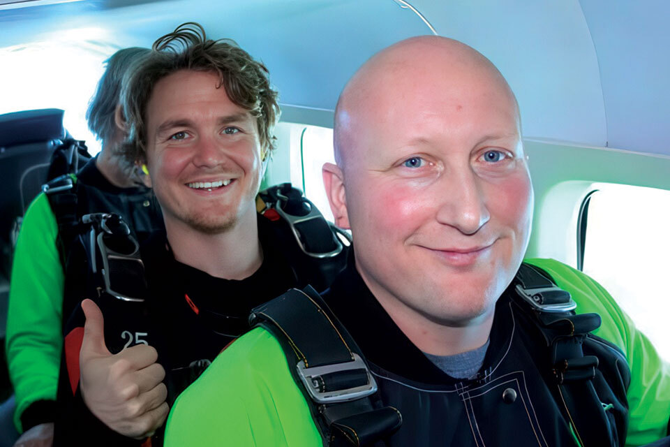 Two men in a plane wearing skydiving gear smile at the camera. The man in front is bald, while the man behind has curly hair and gives a thumbs-up. Both are wearing green and black suits, preparing for a skydive.