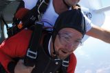 A tandem skydiver with an instructor, wearing helmets and goggles, exits an airplane. The skydiver wears a red suit, and the instructor is in a white shirt. The background shows a clear sky.