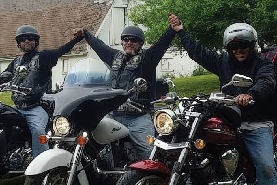 Three motorcyclists in helmets and leather jackets hold hands while sitting on their parked motorcycles. They are outdoors in front of a white building with trees in the background.