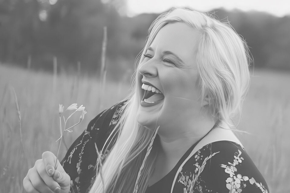 A woman with long blonde hair is laughing joyfully while sitting in a field. She's wearing a floral-patterned dress and holding a small flower. The background is a blurred natural landscape, giving a serene and happy atmosphere.