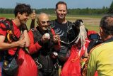 A group of four skydivers, including an elderly man holding a baseball, pose happily on a grassy landing area. One man gives a thumbs up. They are surrounded by parachute gear under a clear blue sky.