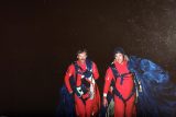 Two skydivers in red suits walk together in the dark, carrying blue parachutes. They appear satisfied and are equipped with various gear.