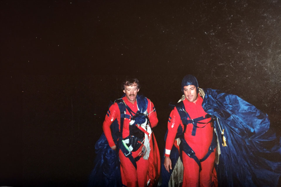 Two skydivers in red suits walk together in the dark, carrying blue parachutes. They appear satisfied and are equipped with various gear.