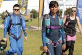 Three people in colorful jumpsuits are smiling and walking on grass. They are carrying helmets and have parachute gear, indicating they are skydivers. Banners and other people can be seen in the background.