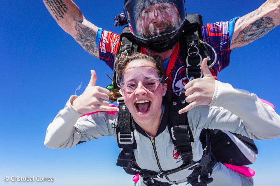 A person tandem skydiving with an instructor. They are smiling, wearing skydiving gear, and making hand gestures against a clear blue sky. The instructor's arms are outstretched.