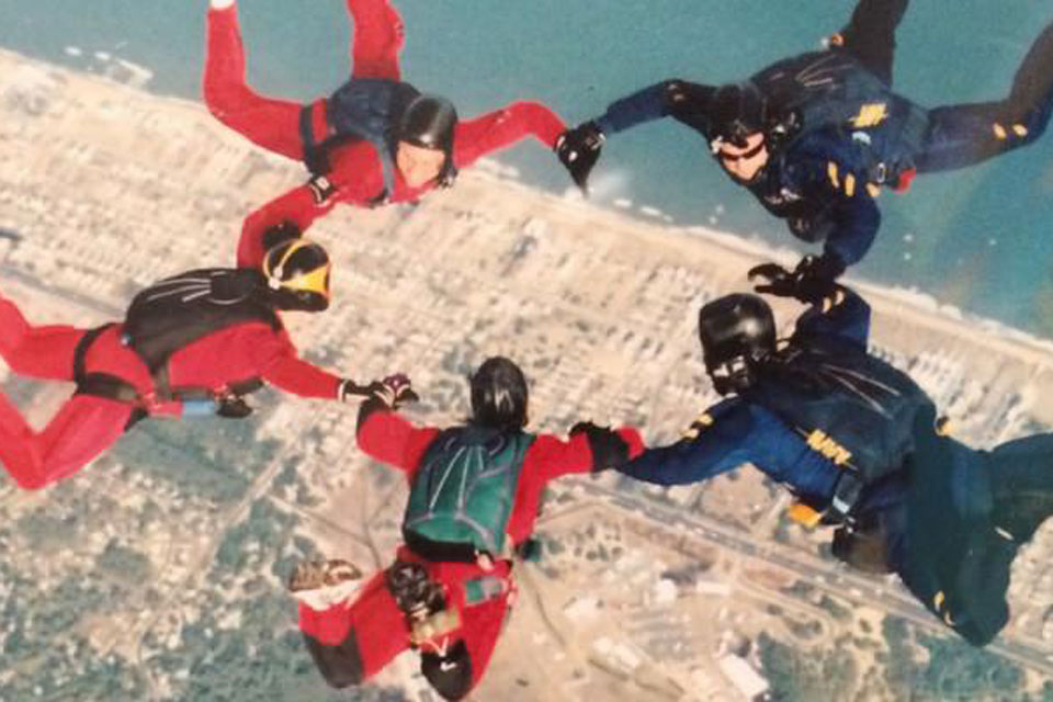 Five skydivers in red and blue jumpsuits form a circle as they freefall above a coastal area, with the sea and land visible below them.