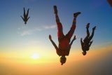 Three skydivers in freefall, with vibrant suits and helmets, perform aerial maneuvers against a stunning sunset backdrop. The sky is a gradient of soft orange and blue hues, adding to the sense of adventure and freedom.