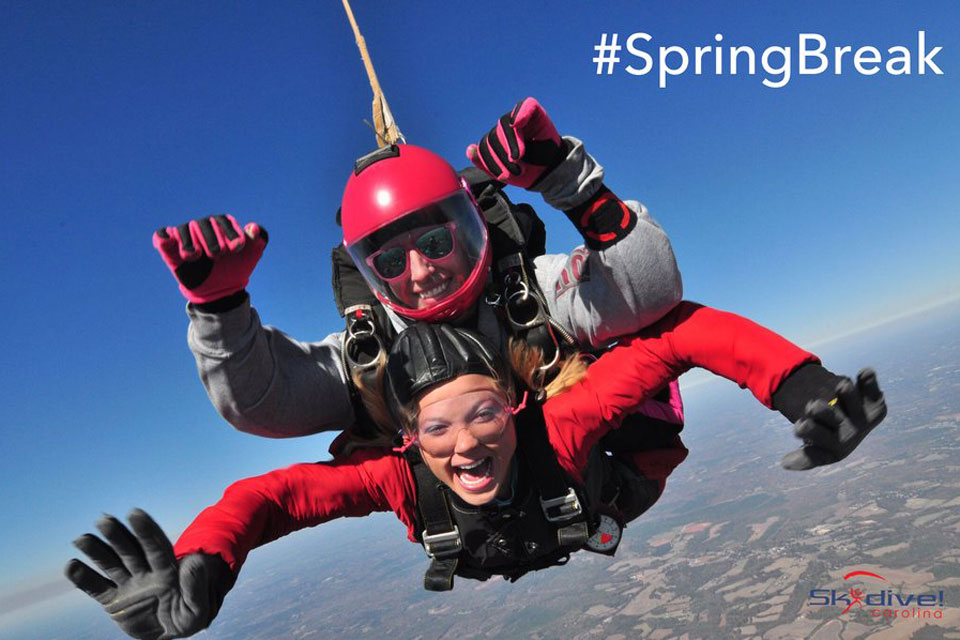 Two people tandem skydiving, both wearing goggles and helmets. The sky is clear blue, and the ground is visible below. The text "#SpringBreak" is in the top right corner, with "Skydive Carolina" logo at the bottom.