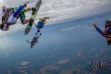 Skydivers in mid-air perform acrobatic maneuvers against a backdrop of open sky and distant landscapes. They wear colorful gear, with one person on a board, showcasing a dynamic and adventurous scene.