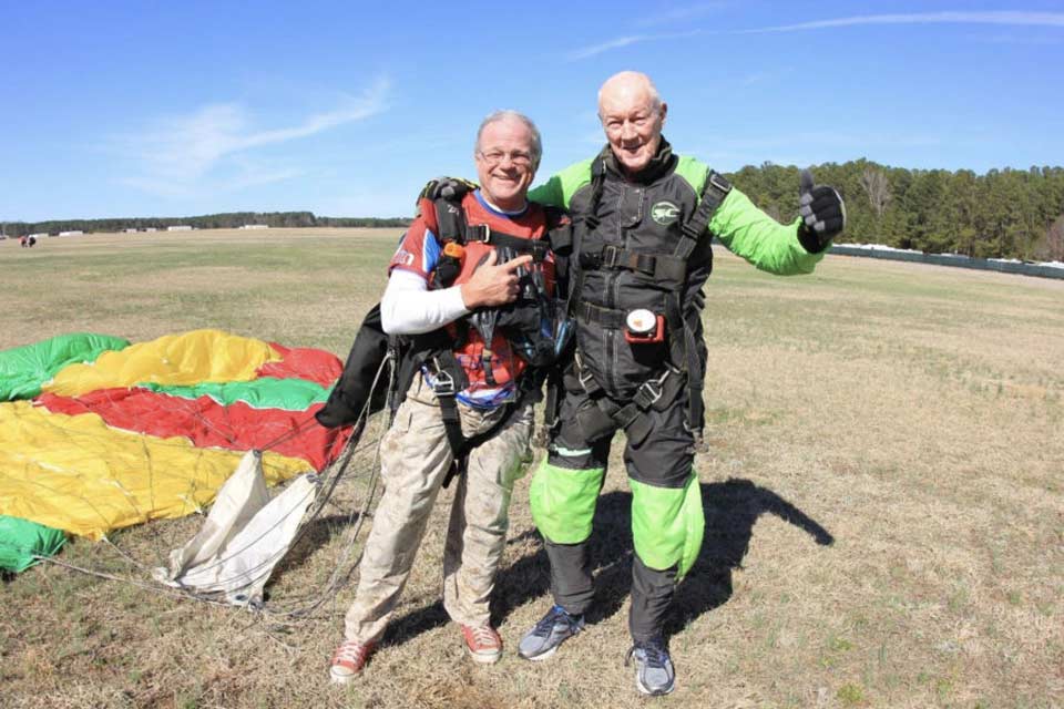 Two smiling men wearing parachuting gear stand on a grassy field beside a colorful parachute. The sky is clear and blue, and trees line the horizon in the background.