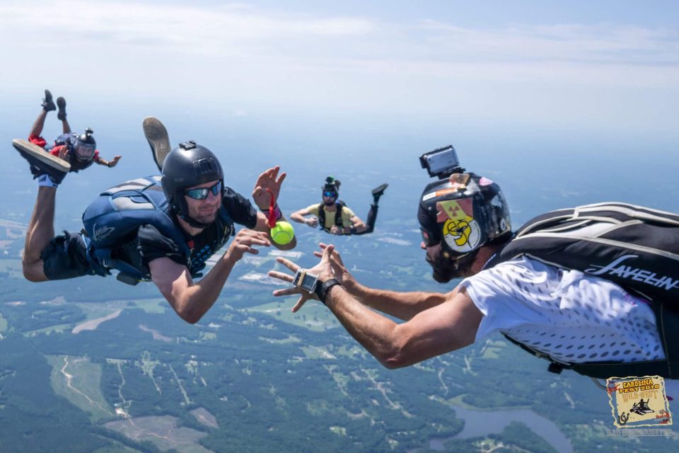 Skydivers in mid-air passing a tennis ball, each wearing helmets and harnesses. Aerial view of a landscape below with fields and forests. The sky is clear and bright, adding to the adventurous atmosphere.