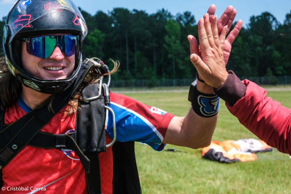 A person wearing a helmet and sunglasses high-fives another individual outdoors, celebrating a successful skydive. A parachute is visible on the grass in the background under a clear blue sky.