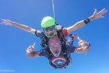 Two people tandem skydiving with clear blue skies. The front person wears a Captain America T-shirt, smiling and giving thumbs up. The person behind wears a green helmet and extends their arms wide. Both have goggles and parachute gear.