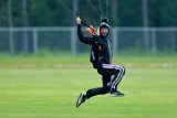 A parachutist in a black jumpsuit and helmet is landing on a grassy field. They are smiling and holding onto the parachute cords. The background is blurred, showing green grass and some trees.